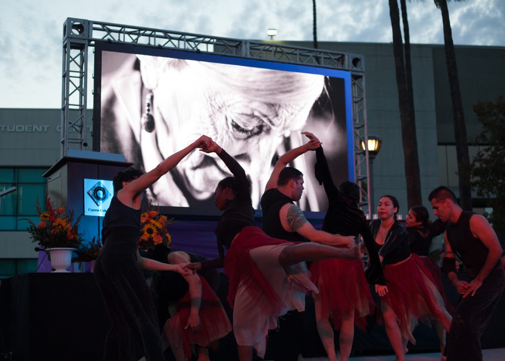 Dancers perform in front of a screen featuring an elderly woman