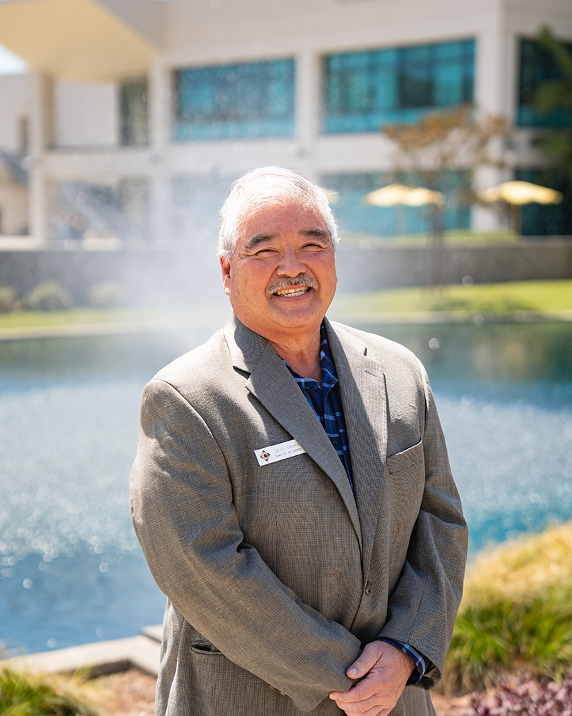 A smiling individual in a gray suit stands outdoors near a body of water, with a modern building and umbrellas visible in the background.
