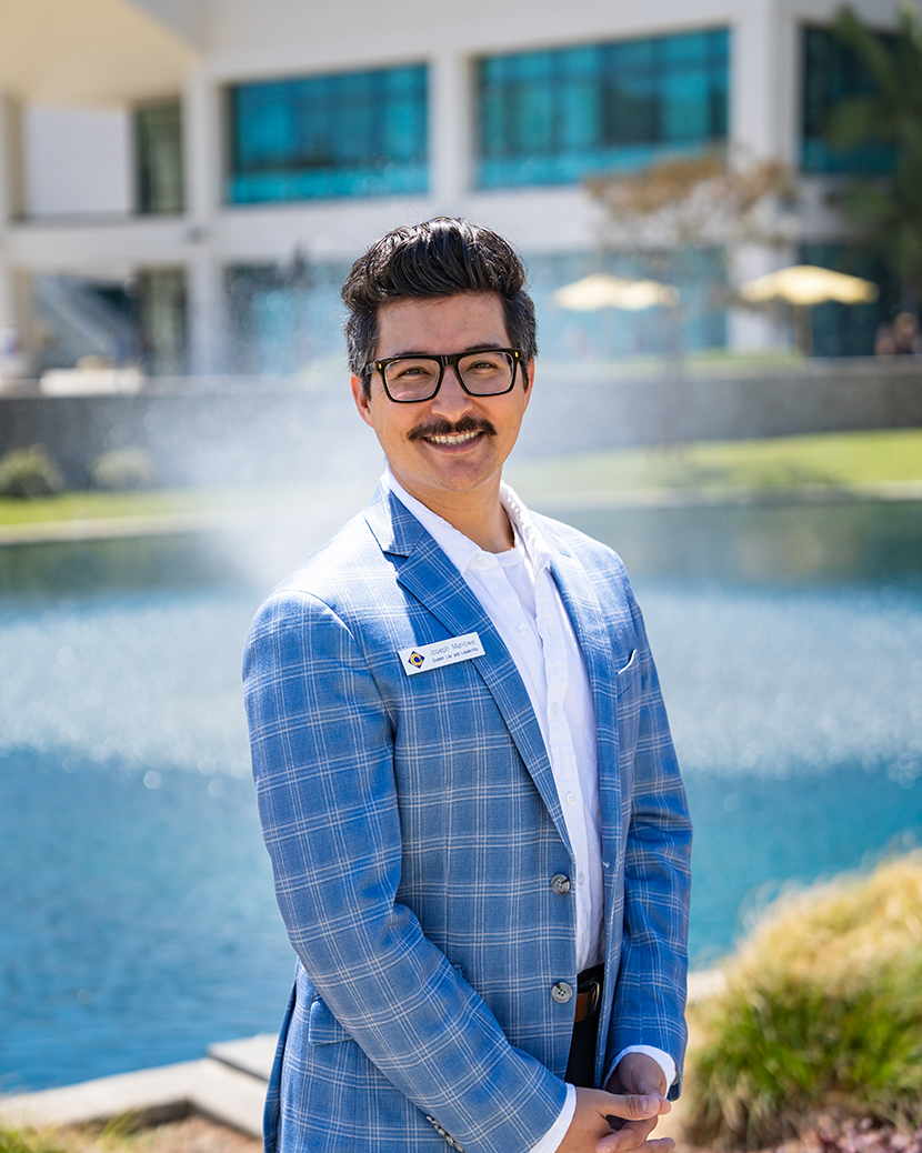 A person wearing glasses and a blue plaid blazer stands smiling by a poolside, with a modern building in the background.