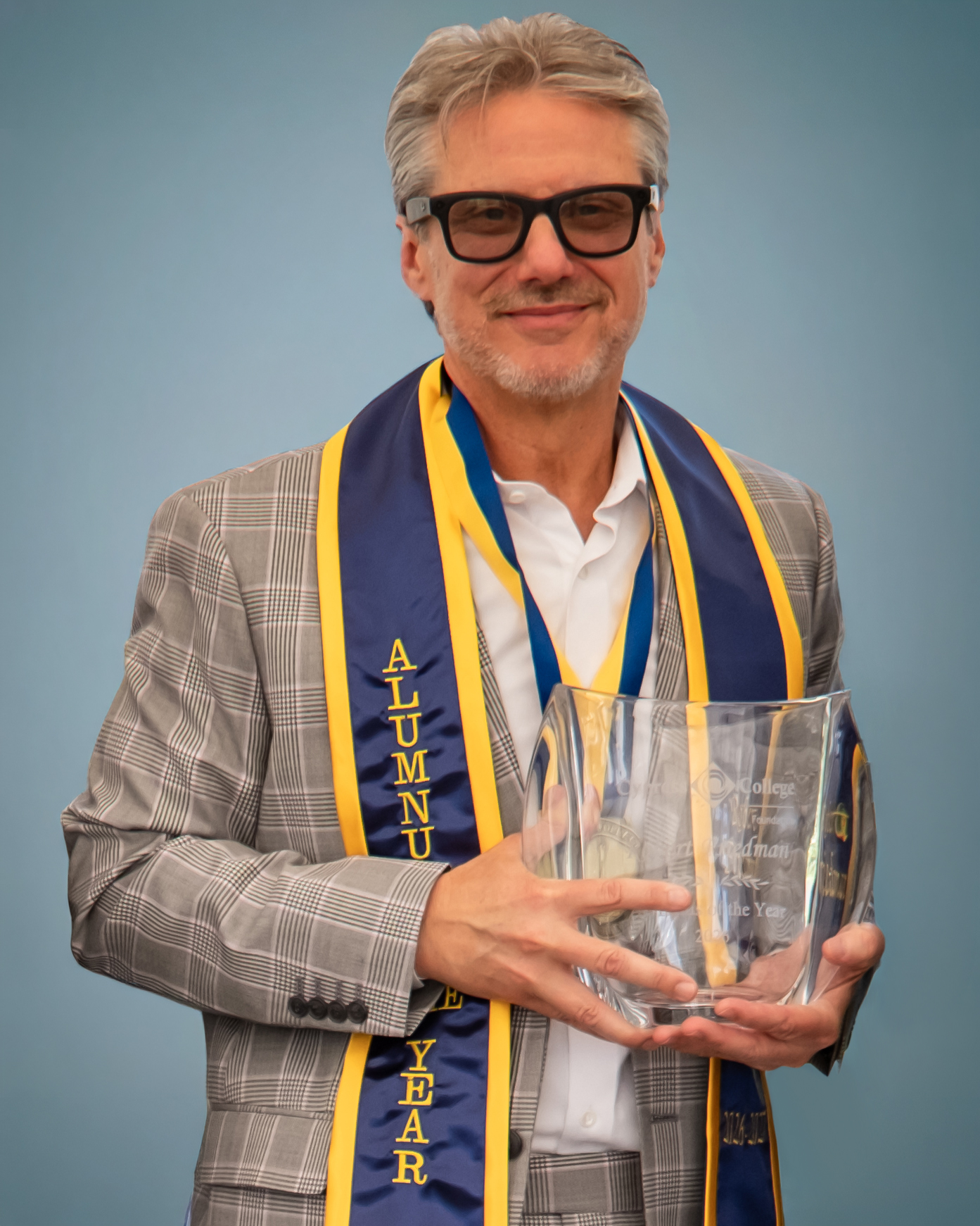 Portrait of Robert Friedman wearing a brown plaid suit, alumnus of the year sash, and holding a trophy