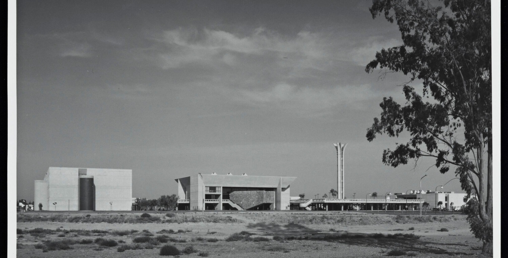 A black-and-white photograph captures a modernist architectural complex with distinct geometric structures set against a vast, open landscape under a cloudy sky.