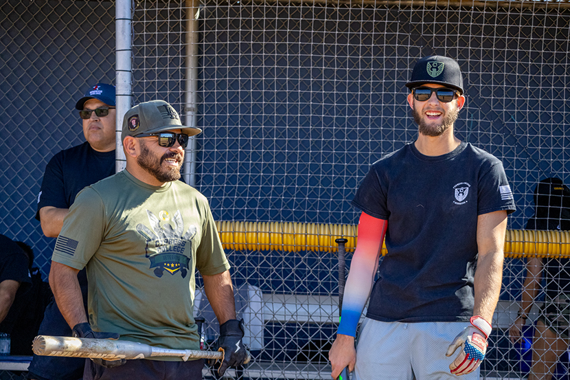 Two men in front of a dugout, one holding a bat and the other smiling.