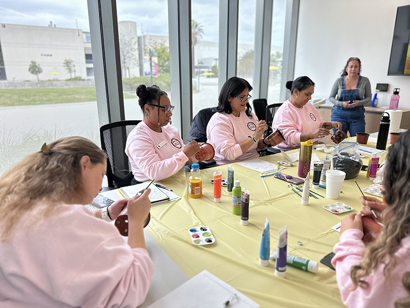 Women Unity event, women are painting pots for plants while someone is presenting. Paint bottles on a covered table.