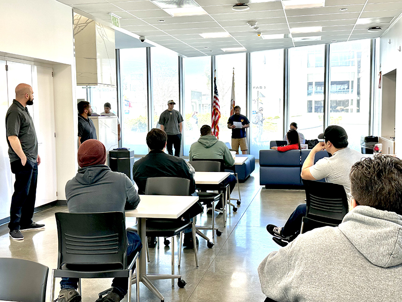 People sitting in a common area, while there is a person presenting In front of tall windows