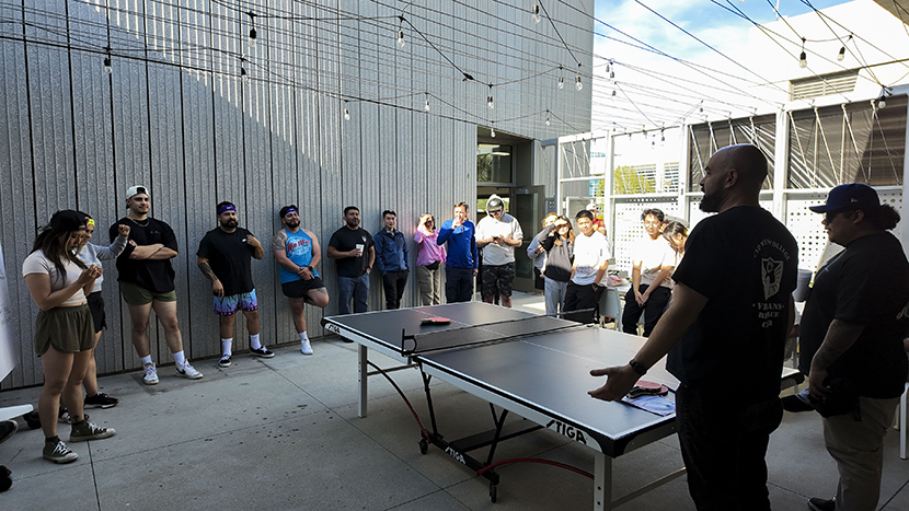 People gathered around a ping pong table, watching someone talking about the rules of ping pong