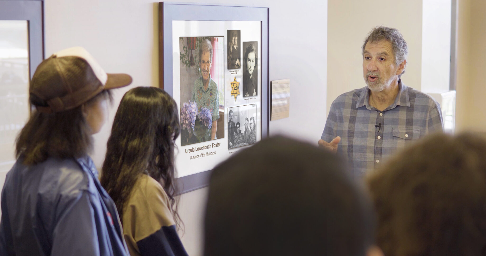 A group of people listens attentively to a speaker in front of a collage of photographs and information about Holocaust survivor Ursula Lowenbach Foster.