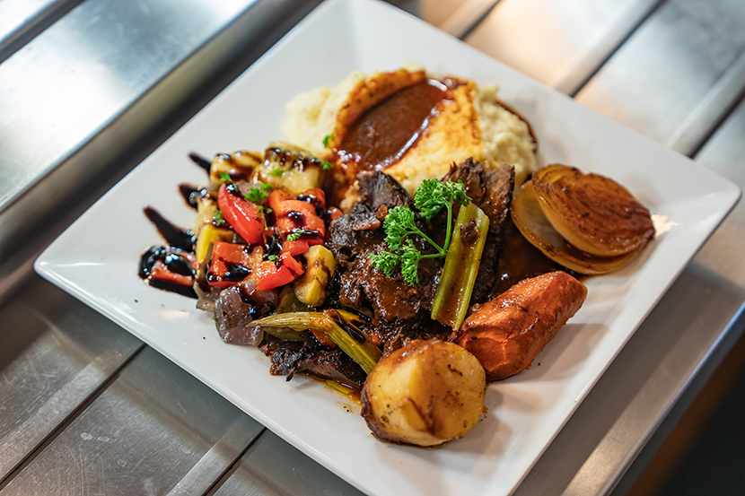 A square white plate with roast beef, mashed potatoes and gravy, glazed vegetables, a roasted onion, and carrots on a metal surface.