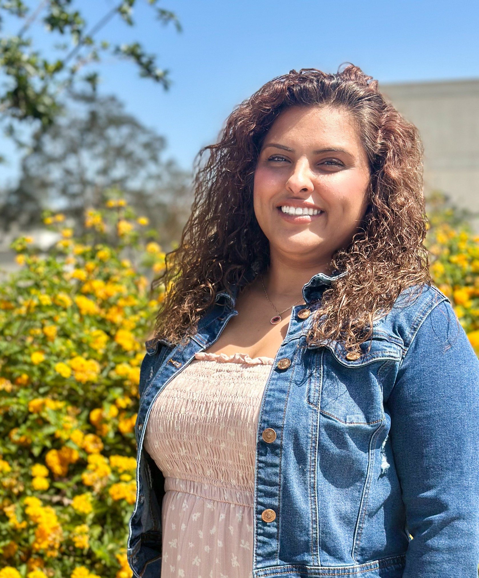 Esmeralda Martin, Adjunct Counselor, standing in front of yellow flowers, wearing a pink smocked dress with a blue jean jacket