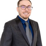 John Risenhoover wearing a dark blazer over a blue shirt standing against a white background, exuding a professional and confident demeanor.