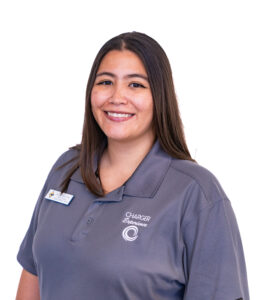 A woman with long straight brown hair and a gray polo shirt smiles at the camera