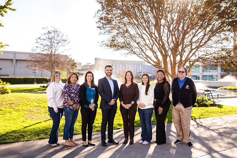 A group of eight adults stands outdoors in a row on a sunny day with trees, grass, and buildings in the background.