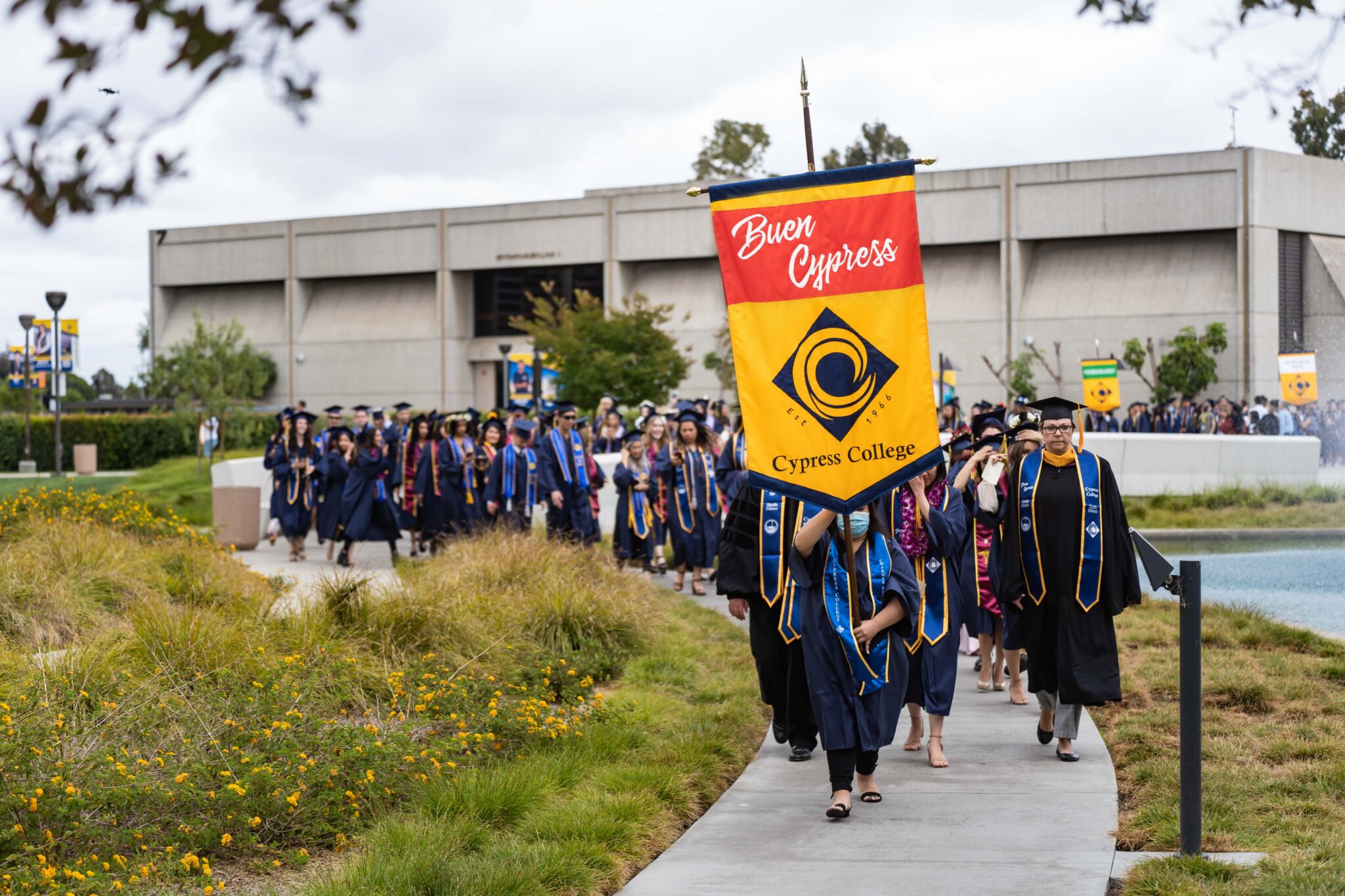 Graduating students walking along path lined with grass near the pond. The person in front is holding a flag that says Buen Cypress and has the Cypress College logo