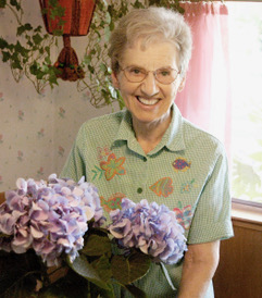 Ursula Lowenbach stands in a bright, floral-themed room, holding a bouquet of vibrant purple hydrangeas.