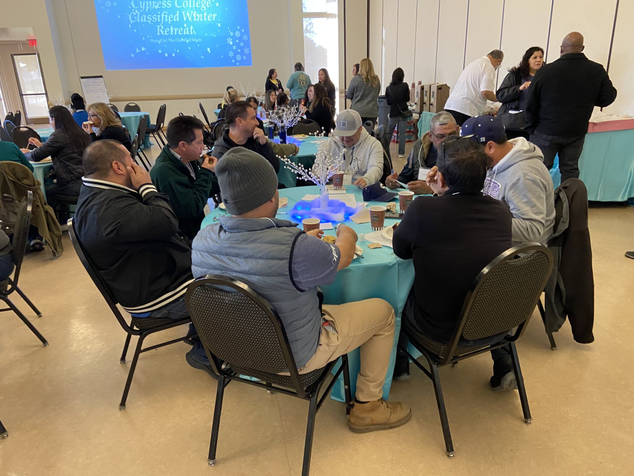 A group of classified staff sitting at a round table and others standing around socializing.