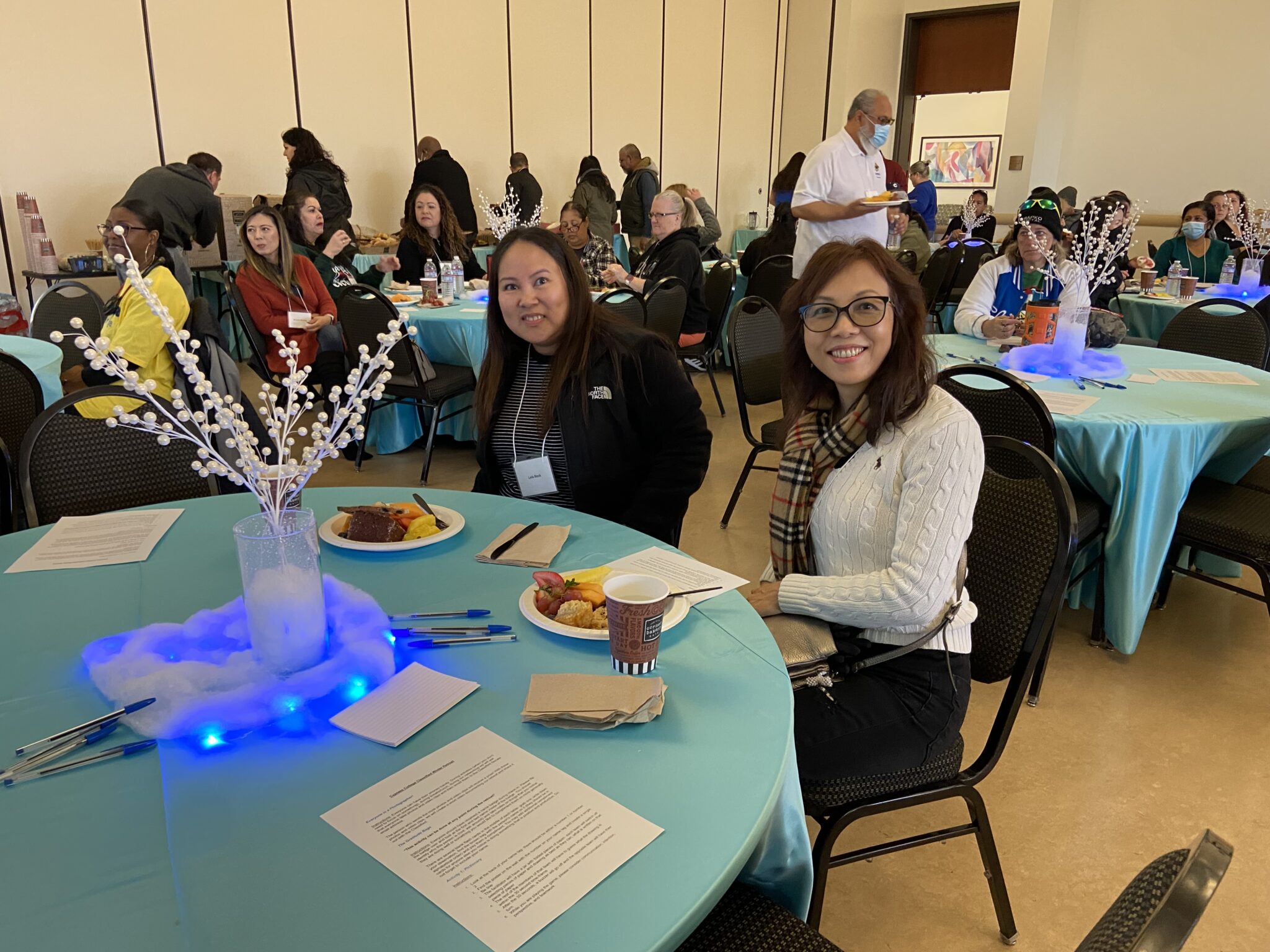 Two classified staff are smiling as they take a photo while sitting at their table and enjoying their breakfast pastries.