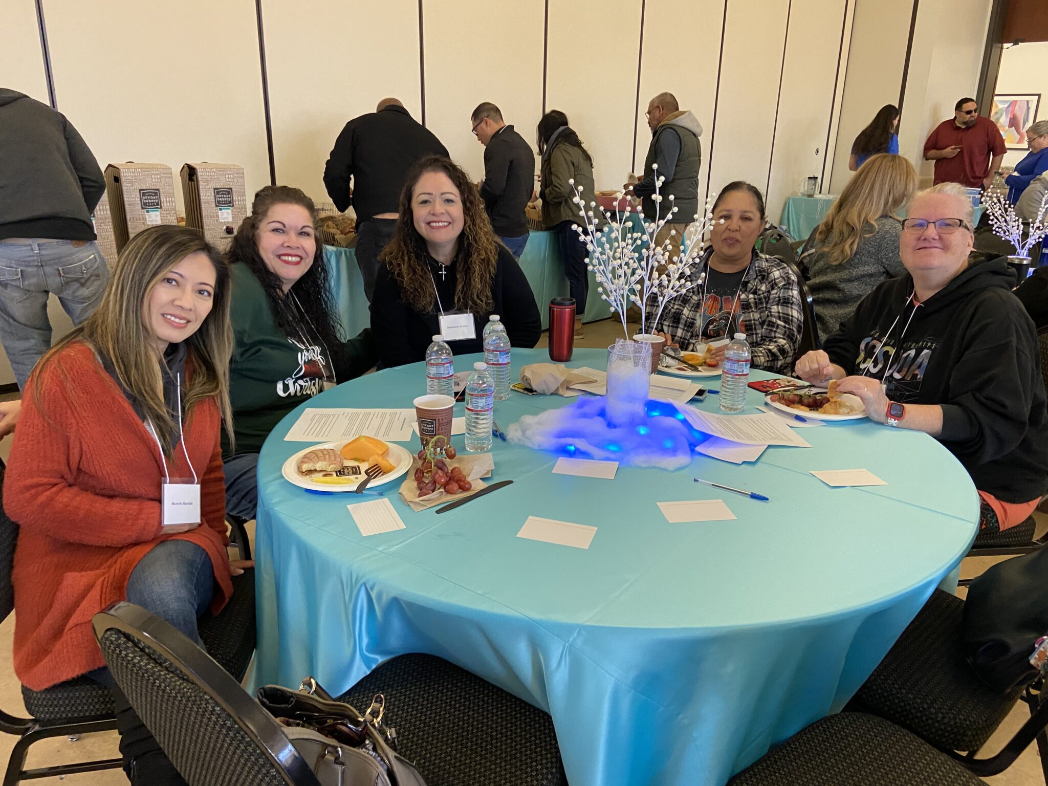 A group of classified staff sitting at a table and posing for a photo while others serve themselves food in the background.