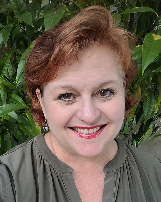 A person (Professor Adrienne Merlo) with short, curly red hair stands in front of lush green plants, wearing a loose olive green top.