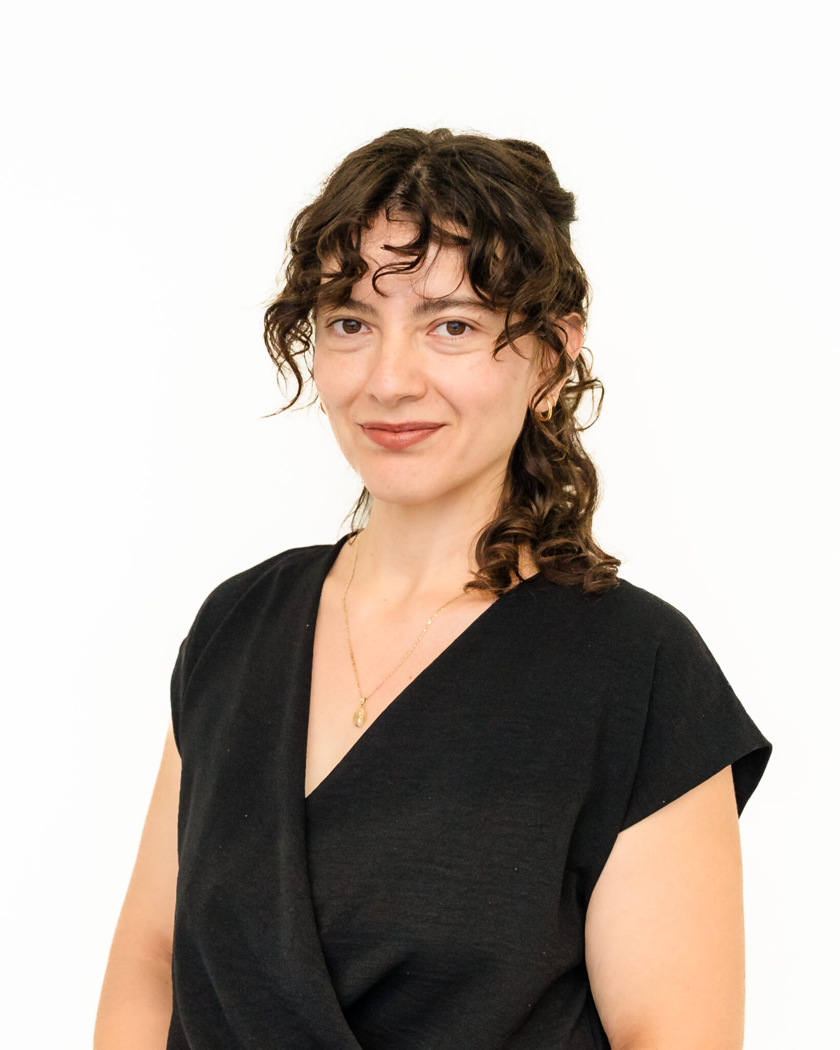Portrait of a smiling woman with curly hair, wearing a black top and a gold necklace, set against a white background.