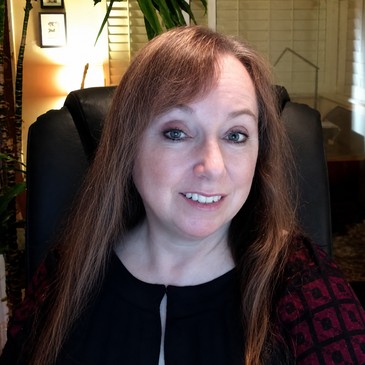 Smiling woman with long brown hair sitting in a black chair in a warmly lit room with plants and framed pictures.