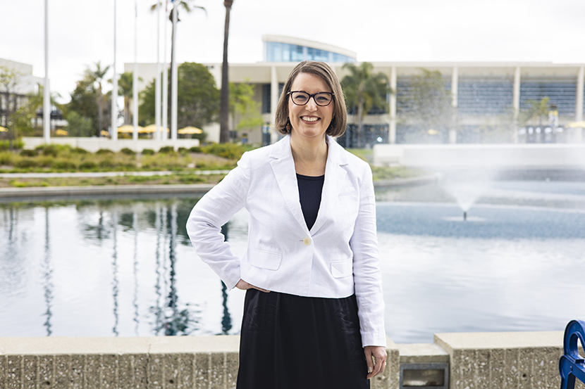 Professional headshot of Jill Bauer smiling in glasses and a white coat over a black top, with a campus background.