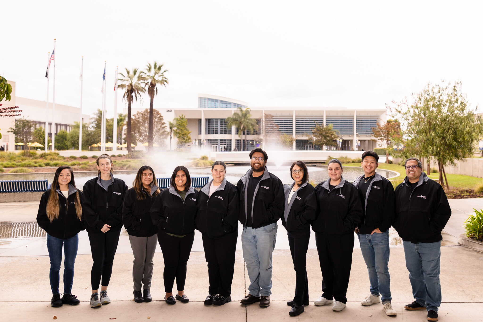 A group of eleven people stands outdoors near a fountain, wearing matching black jackets, with palm trees and a building in the background.
