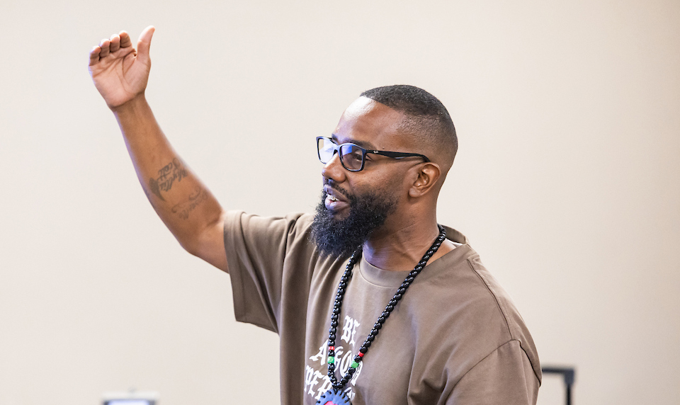 A dark-skinned man with glasses and beard points in an upward motion.