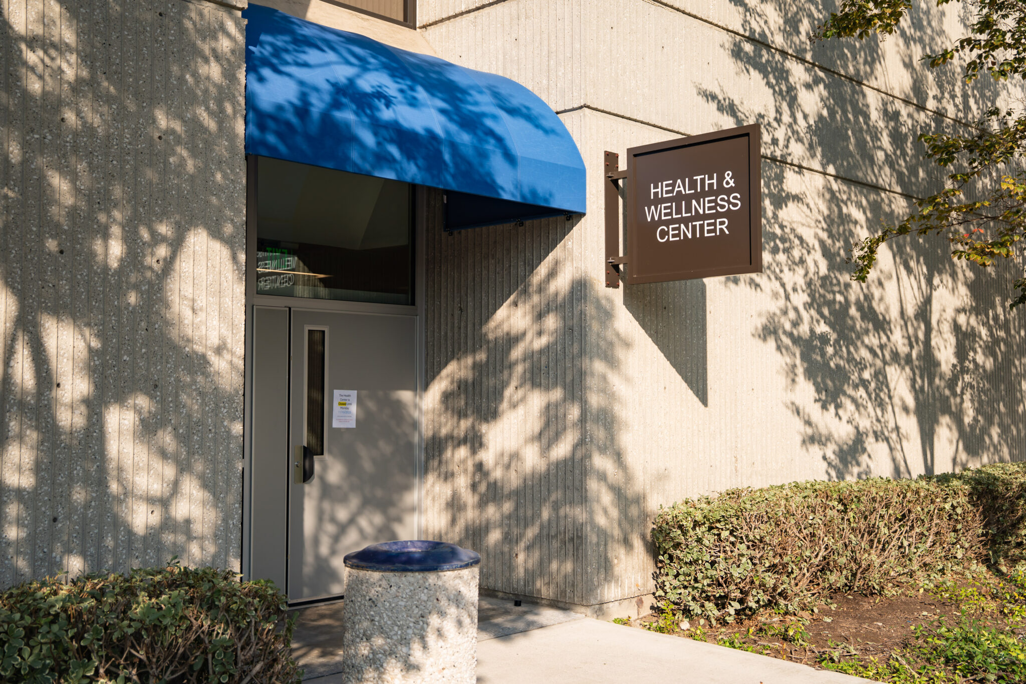 Entrance of the Health & Wellness Center, featuring a blue awning and a sign, surrounded by greenery and concrete walls.