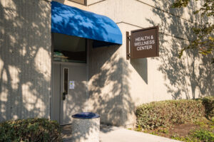 Entrance of the Health & Wellness Center, featuring a blue awning and a sign, surrounded by greenery and concrete walls.