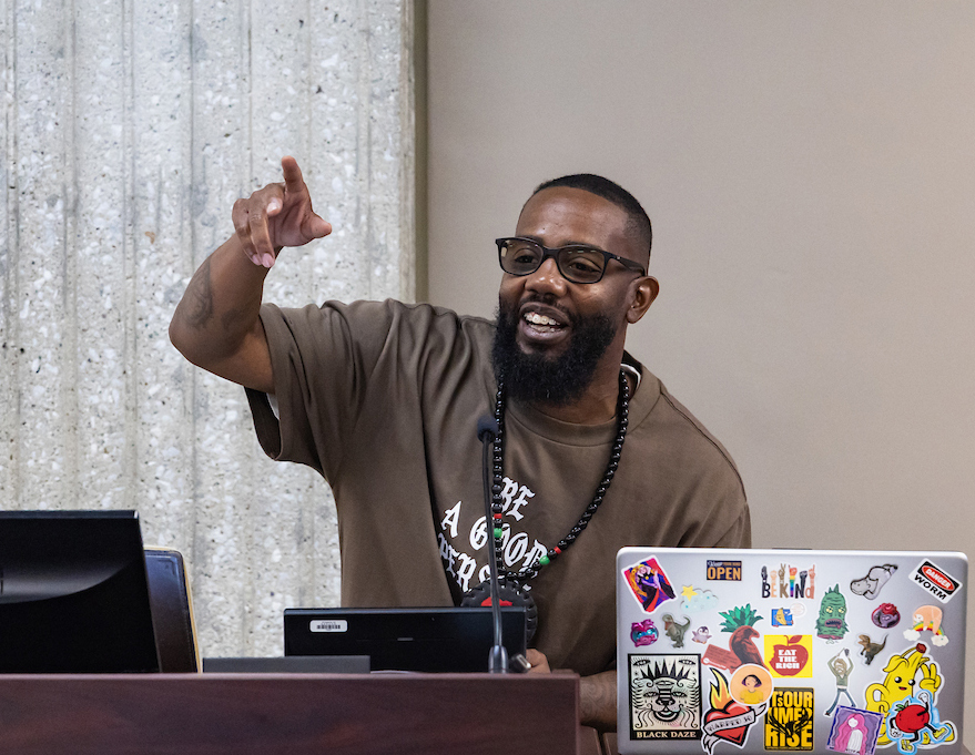 A Black man stands at a podium with a laptop and smiles and points out to an audience.