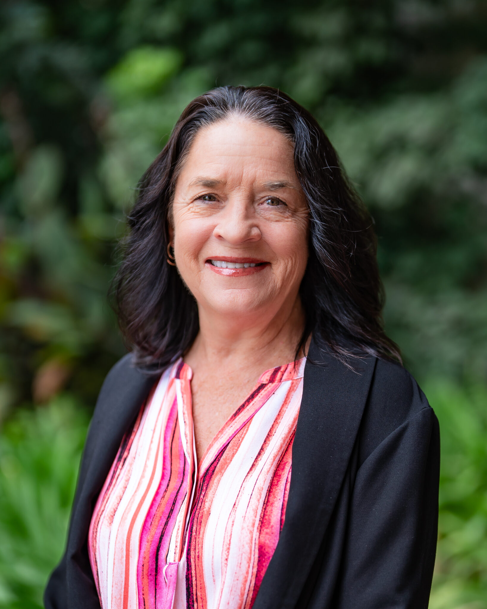 Smiling woman with long dark hair wearing a pink-striped blouse and black blazer. She stands outdoors with green foliage in the blurred background.