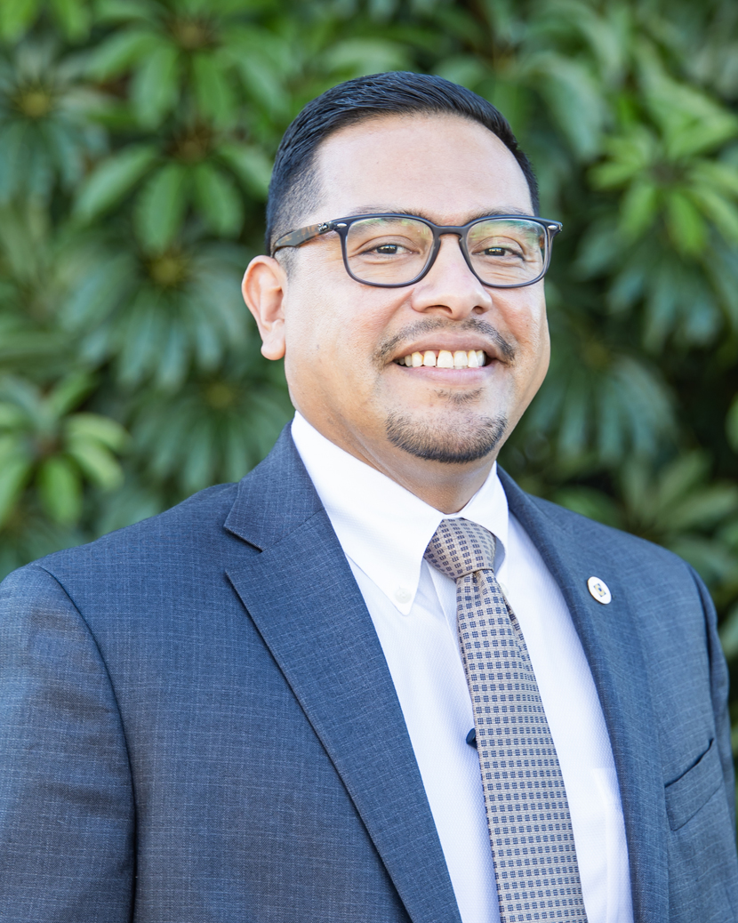 A person, Luis Gonzalez, in a gray suit and patterned tie stands confidently outdoors, surrounded by lush green foliage.
