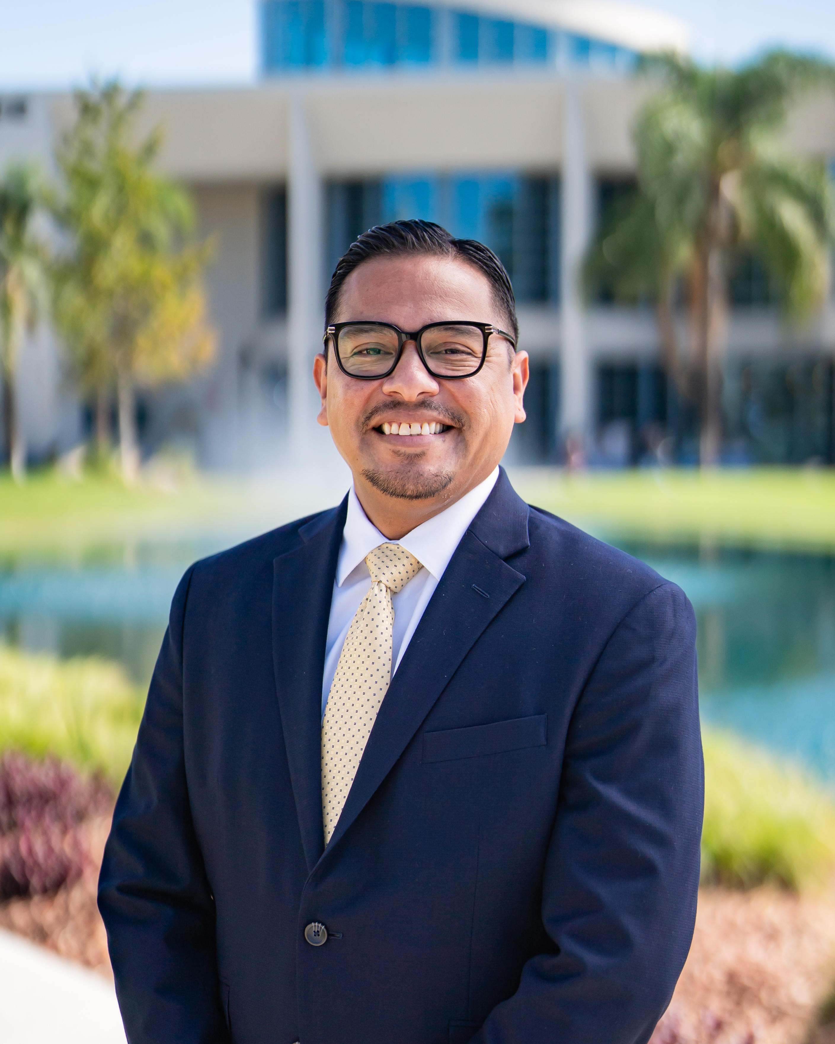 A person, Luis Gonzalez, in a gray suit and patterned tie stands confidently outdoors, surrounded by lush green foliage.