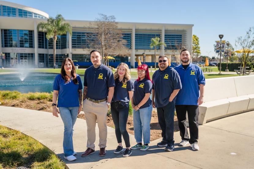 A group of six people wearing matching navy blue shirts with yellow logos pose in front of a modern campus building with large glass windows. They are standing on a pathway near a fountain and palm trees, smiling at the camera.