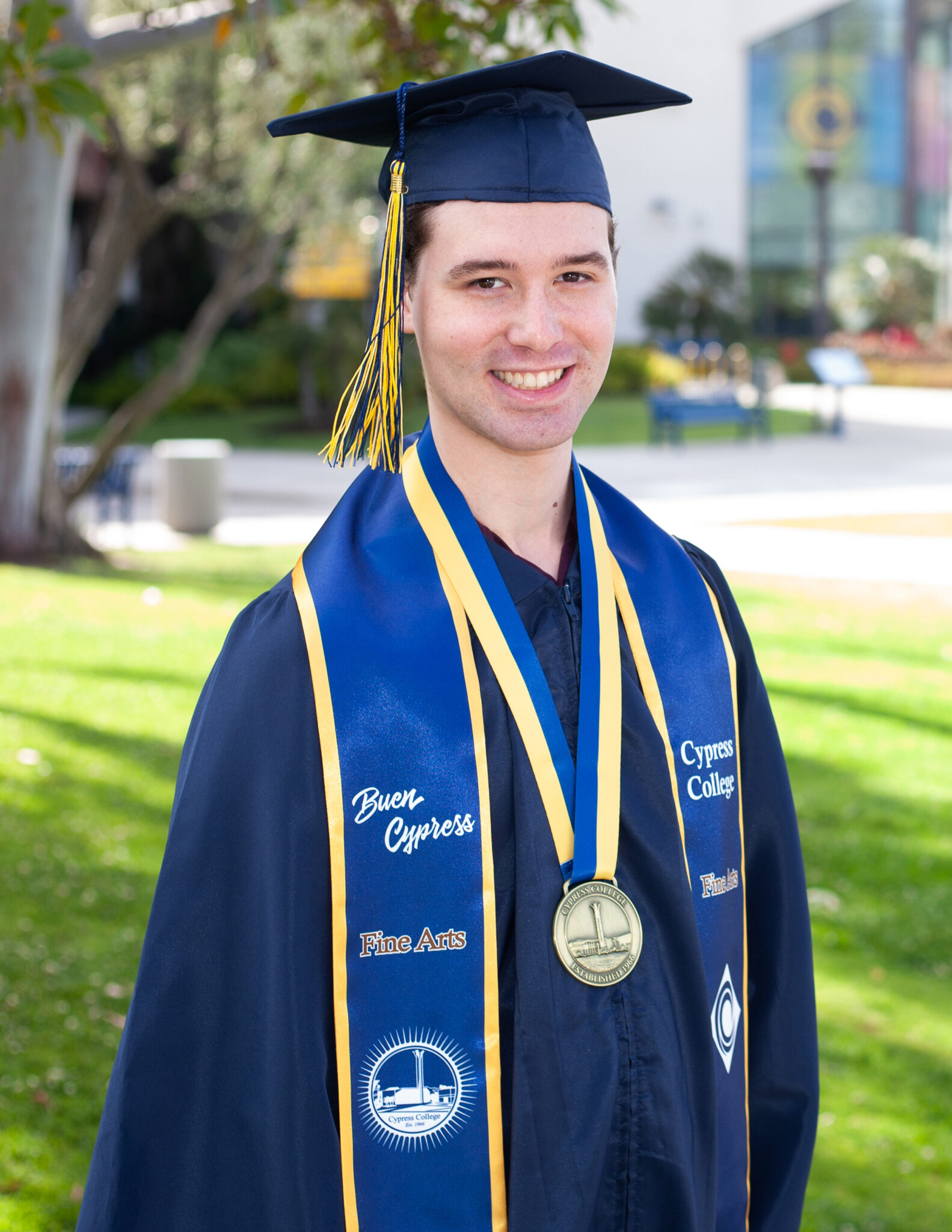 Portrait of Preston Harris, the 2023 Presidential Scholar of Distinction for the Fine Arts pathway, wearing regalia and holding a Cypress College "We Journey Together" pennant with Gateway Plaza, including the Student Center and Campanile, in the background.