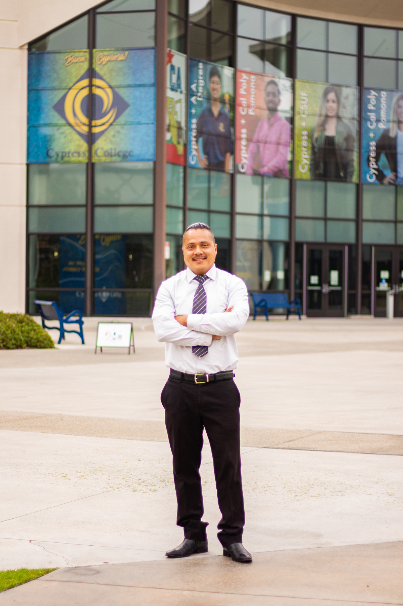 David Garcia, a man wearing dark pants and a white button-down shirt and tie, folds his arms and stands in front of a Cypress College building with banners promoting students.