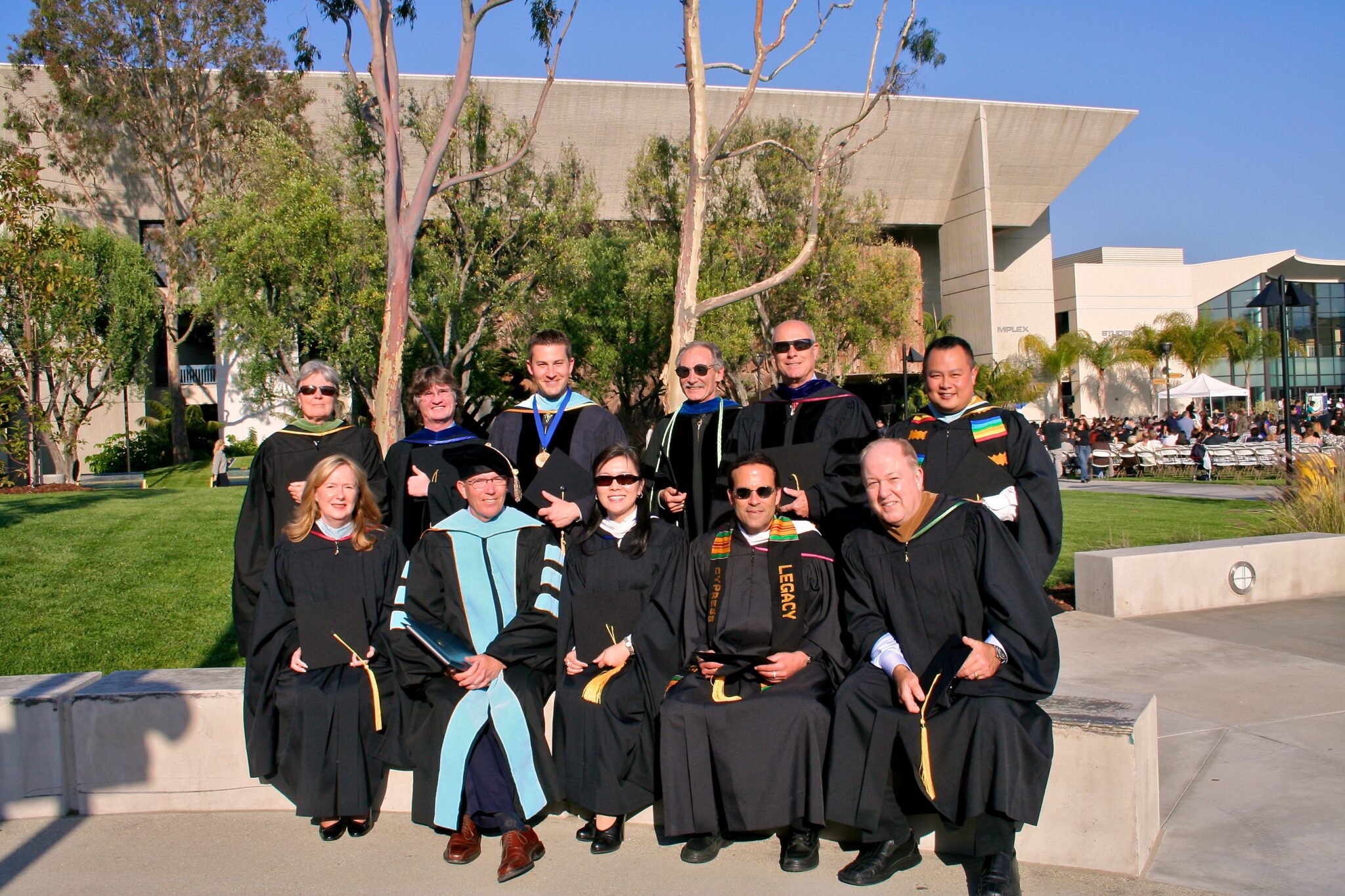 Group of employees in caps and gowns posing outdoors, celebrating commencement with trees and a modern building in the background.