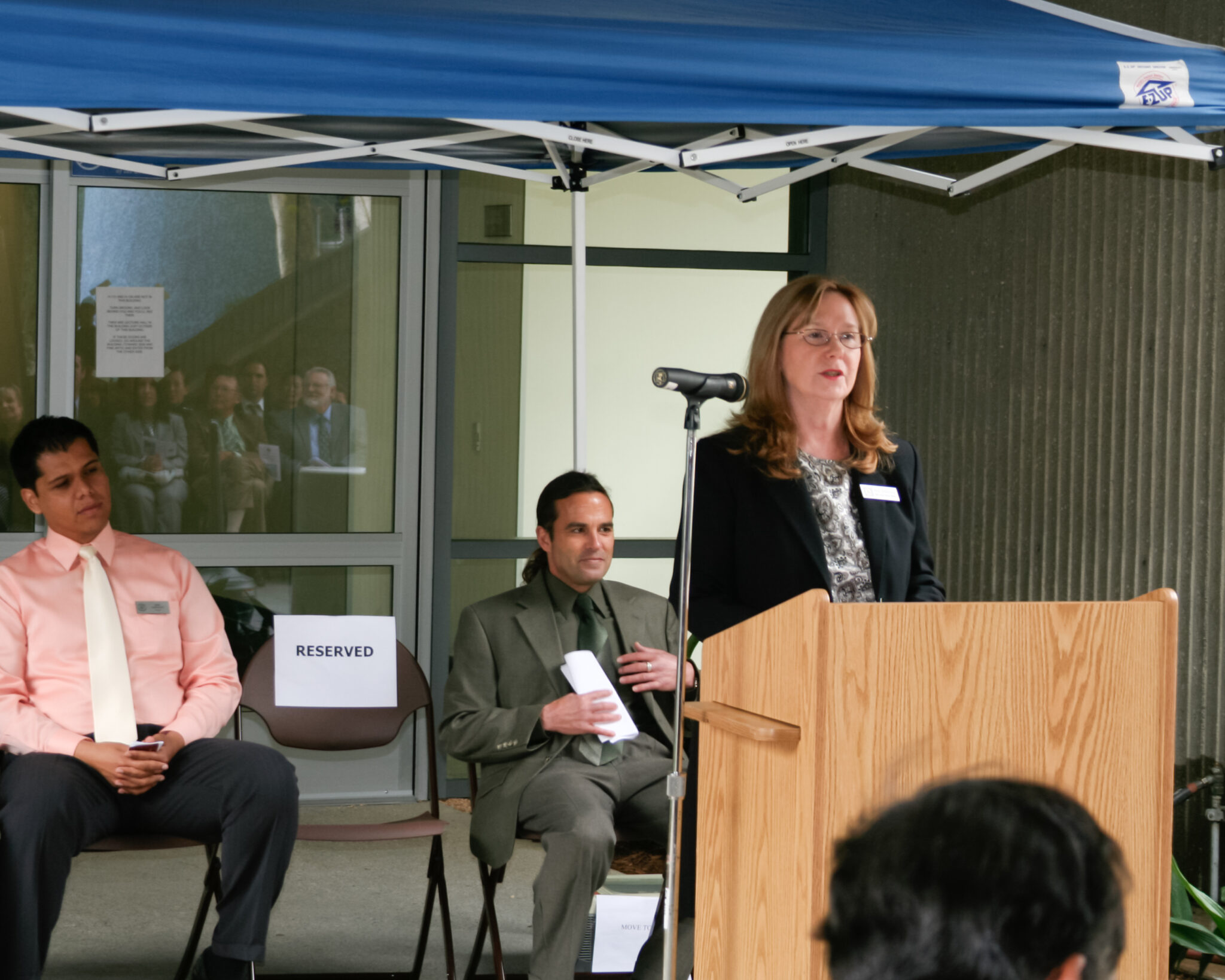A woman, Nina Demarkey, speaks at a podium under a tent, with seated attendees in the background, including one chair marked "RESERVED."