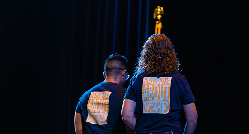 Man and woman with backs turned wearing t-shirts that read 'ask me about accreditation'