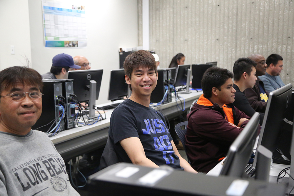 Students sit in front of computers and smile for the camera in a classroom.