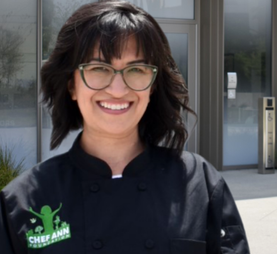 A woman in glasses and a black chef's jacket smiles at the camera.