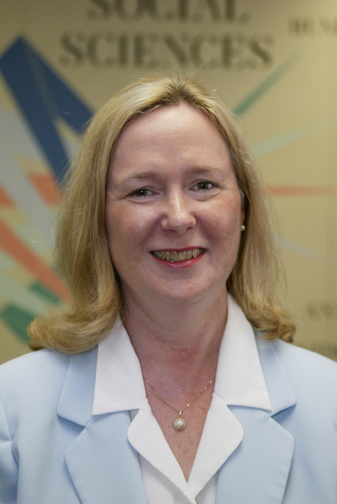 Portrait of Nina DeMarkey in a light blue blazer and white blouse, standing in front of a colorful background related to social sciences.