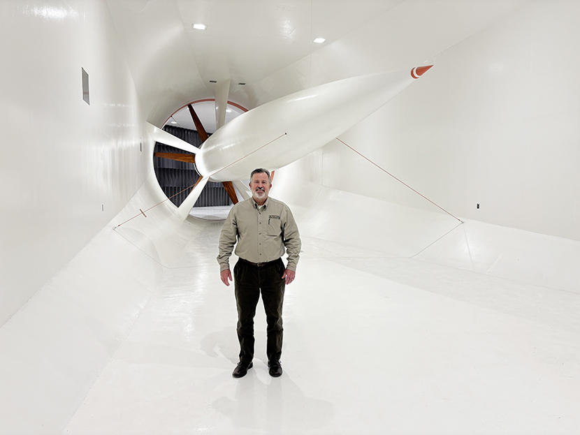 Man standing inside a large white wind tunnel with a wind turbine blade suspended above him.