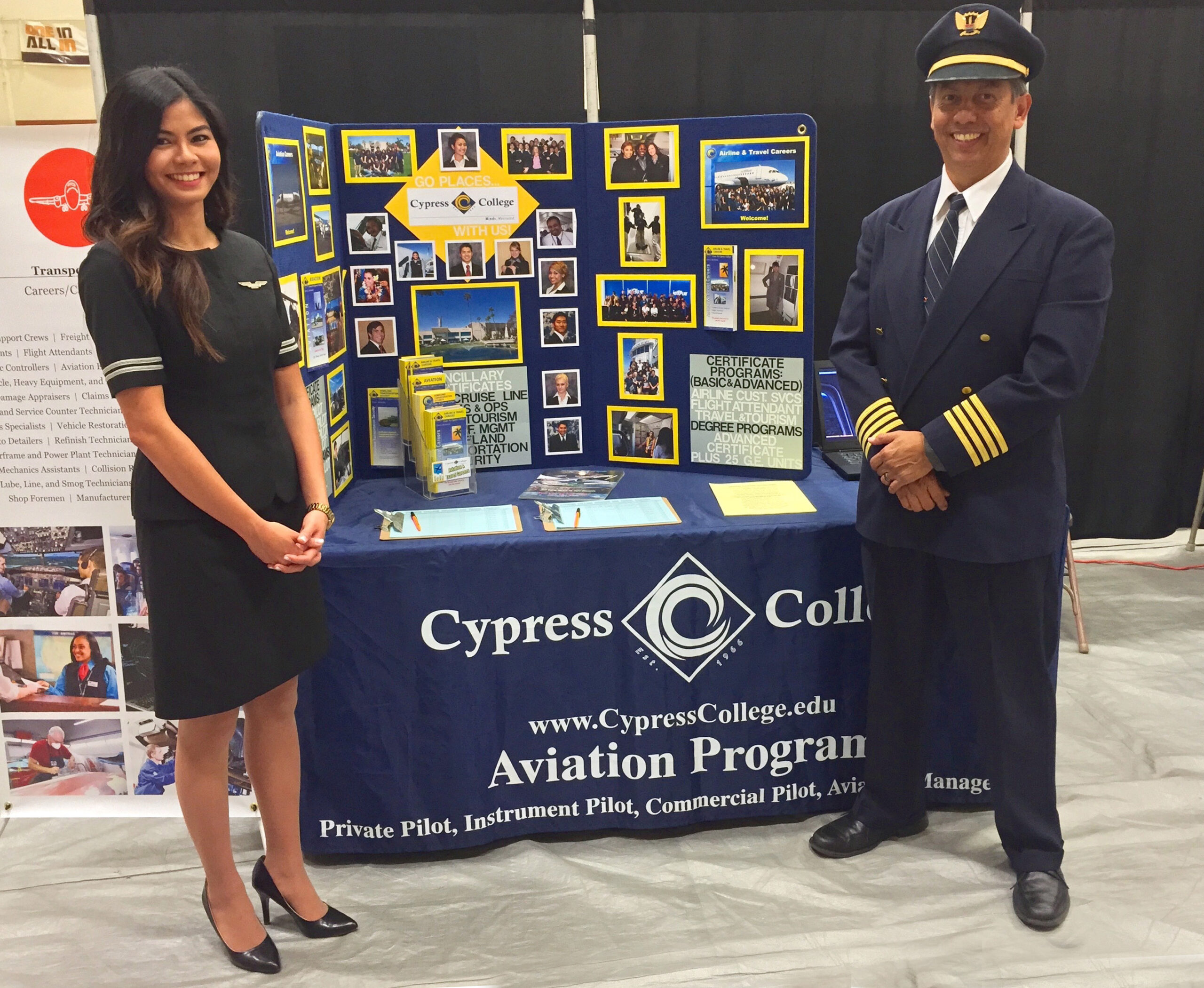 A flight attendant and pilot in navy blue uniforms stand before a table with handouts on information for studying and getting jobs in the aviation field.