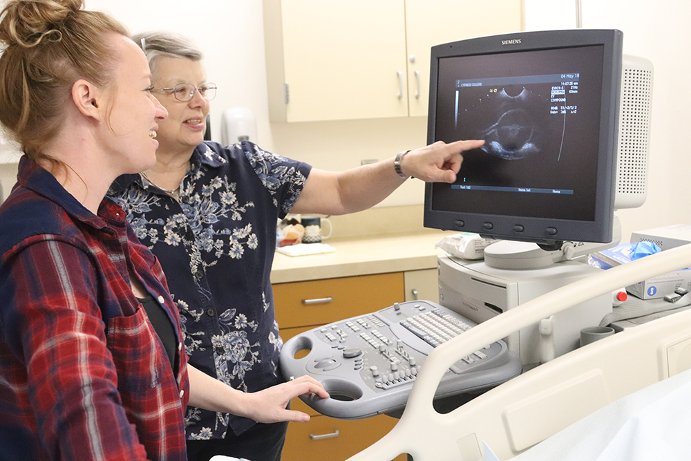 An older woman points to a monitor connected to a sonography tool. A younger woman looks on and smiles.