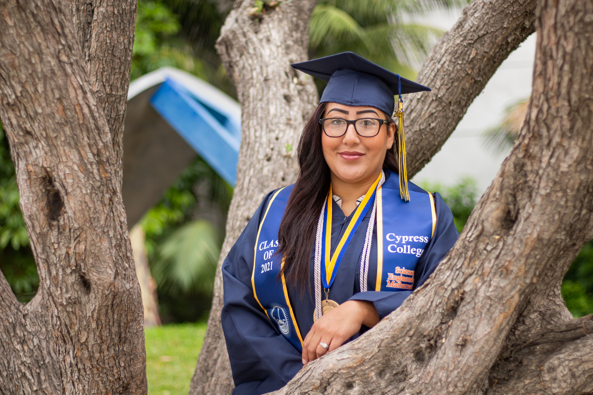 CYProud Student Melissa Whitewater wears graduation regalia and stands by a tree on the Cypress College campus.