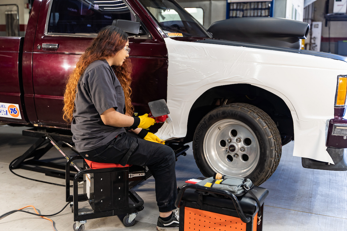 A young woman with long dark curly hair works on the paint job of a boxy car.