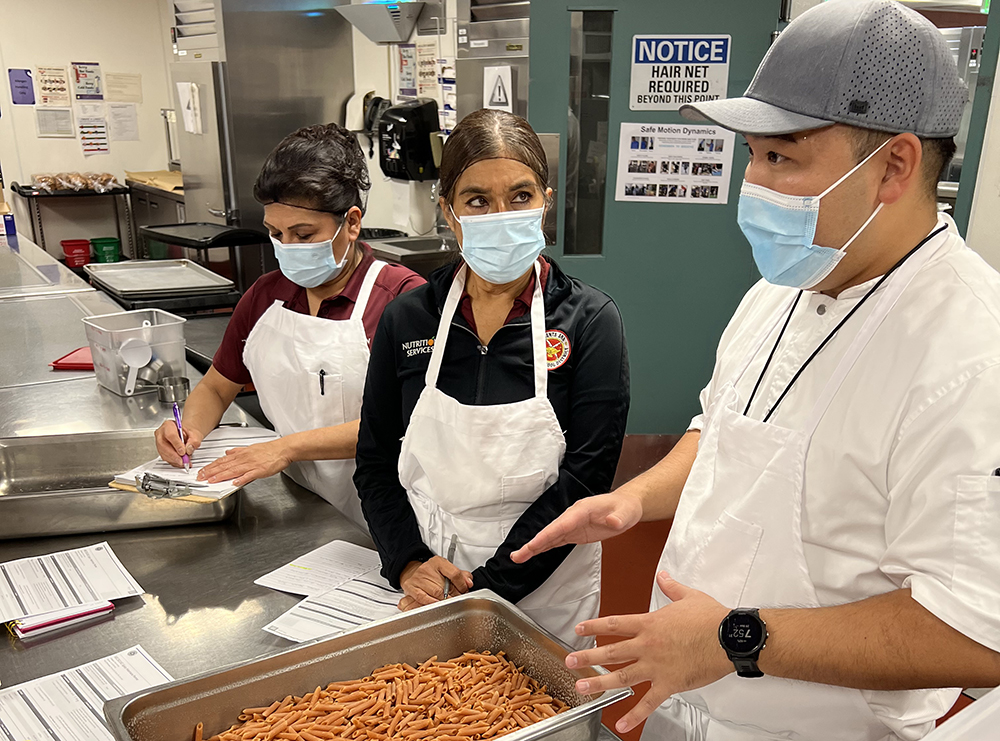 Jonathan Park talks to two lunch cooks in a school kitchen as they prepare pasta.