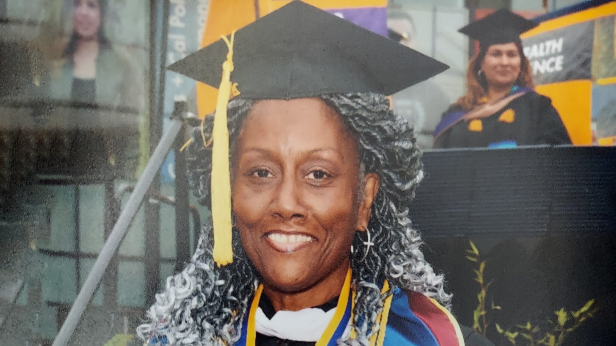 A woman with gray braids and a mortarboard poses in graduation regalia.
