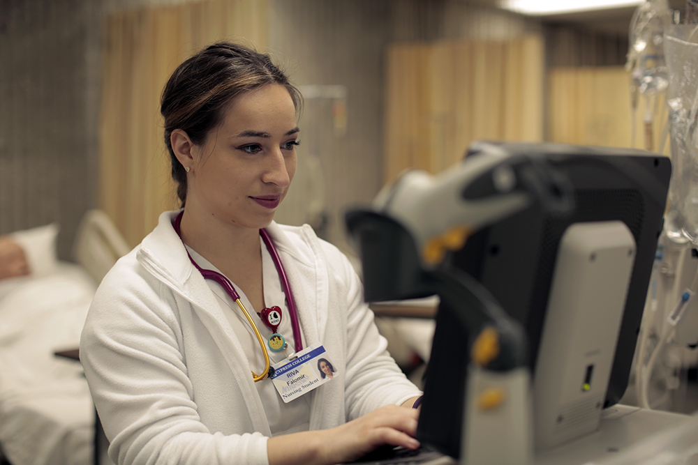 A woman in a nurse's uniform works at a computer in a hospital room setting.