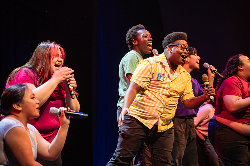 A group of singers performing on stage, holding microphones, with colorful outfits against a dark backdrop.